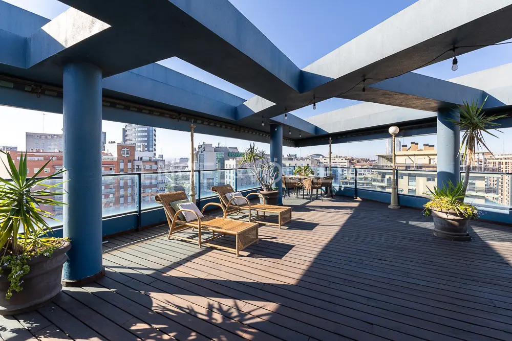 Rooftop patio with blue pergola, wicker furniture, and potted plants. City buildings are visible in the background.