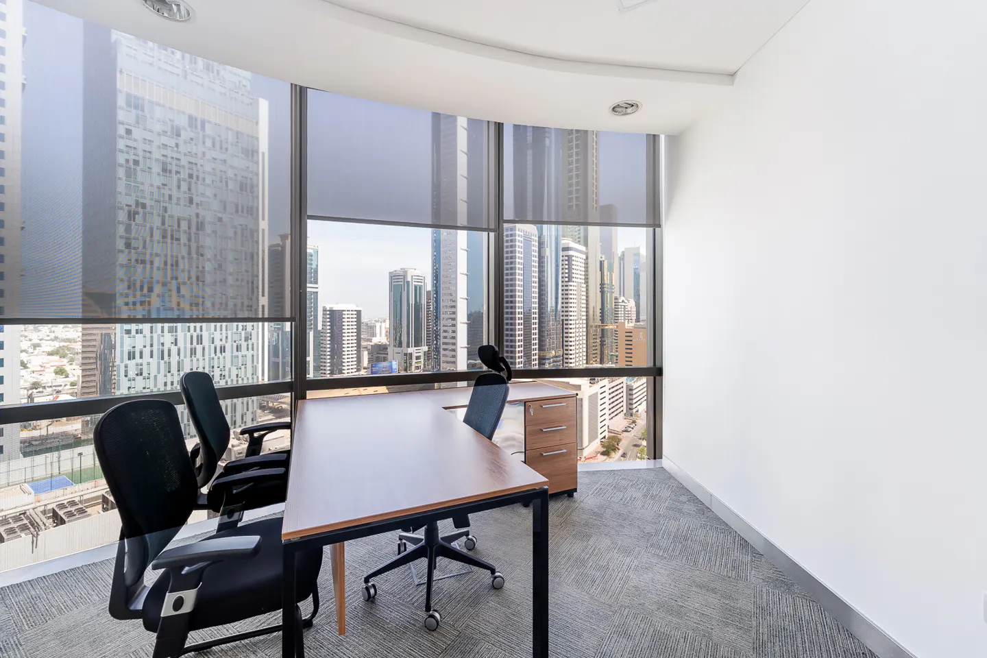 Office space with a desk, chairs, and a view of a city skyline through large windows with gray blinds.