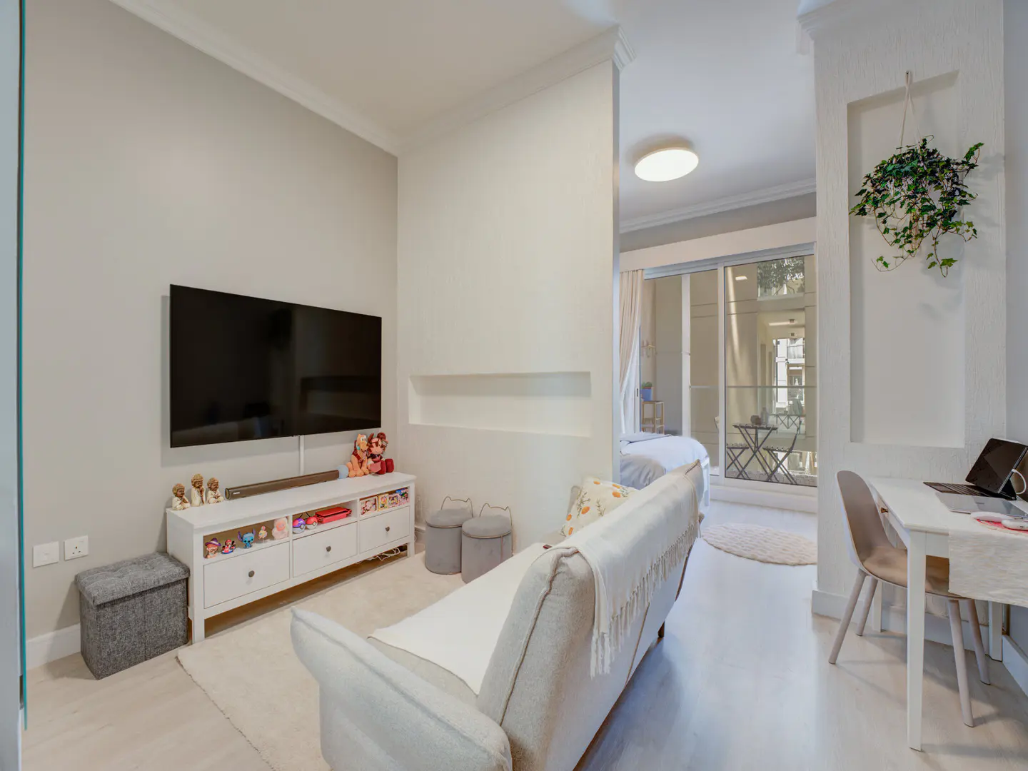 Living room with a TV, white media console, and a gray sofa. A desk and chair are visible in the adjacent room.