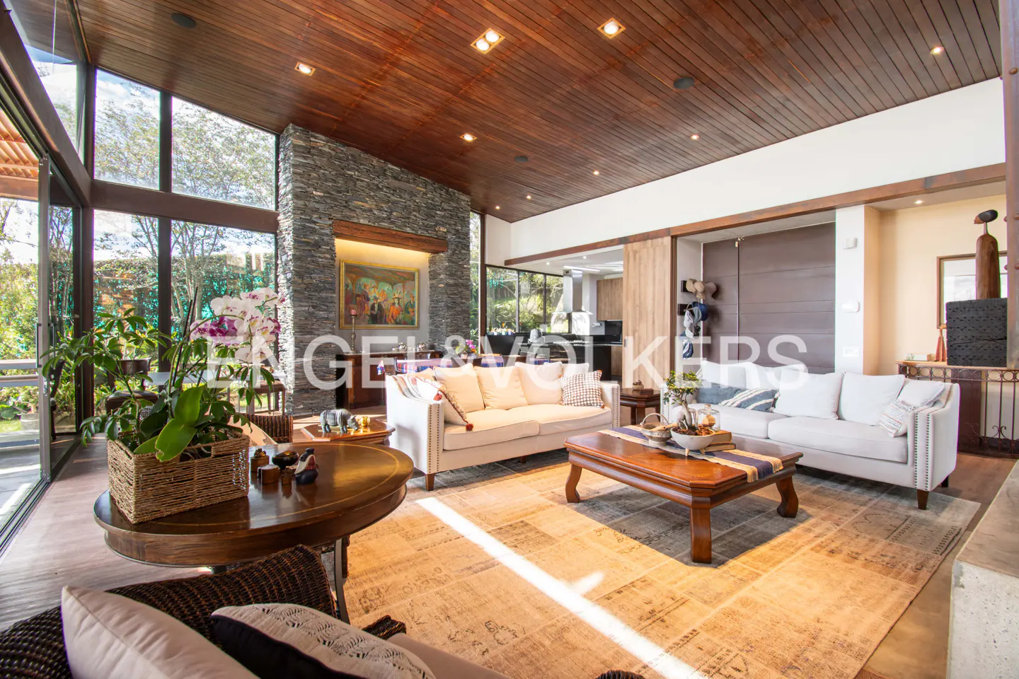 Bright living room with wood ceiling, stone wall, and large windows. Two white sofas face a wooden coffee table on a patterned rug.