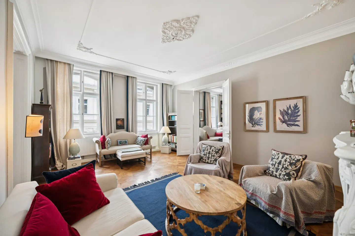 A bright living room with white sofas, red pillows, and a blue rug. A wooden coffee table sits in the center. Two framed leaf prints hang on the wall.