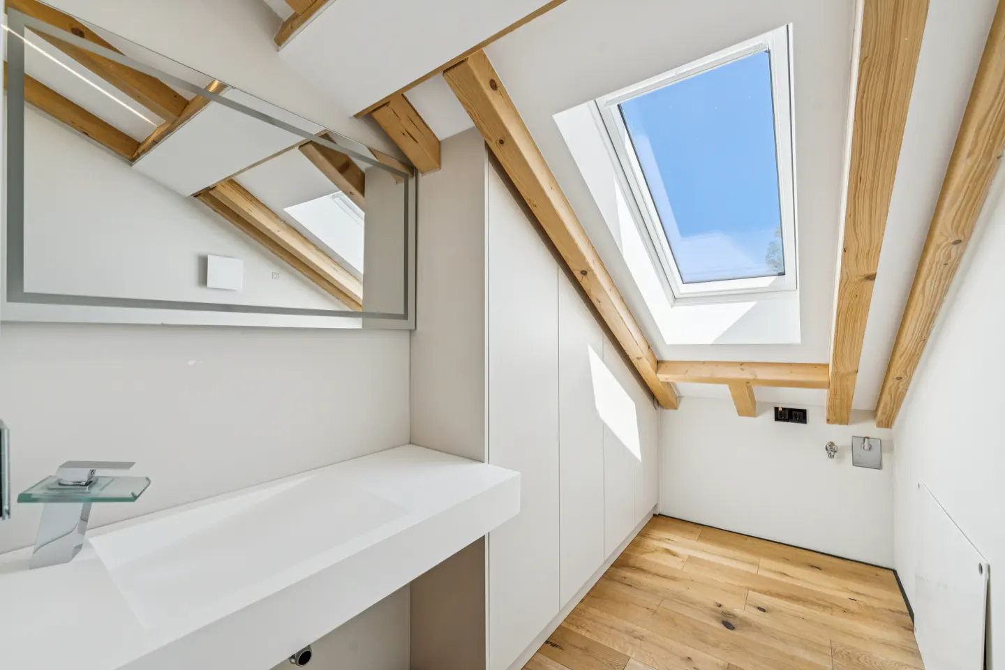 Attic bathroom with white walls, wood floors, and exposed beams. A skylight lets in natural light. A modern sink and mirror are visible.