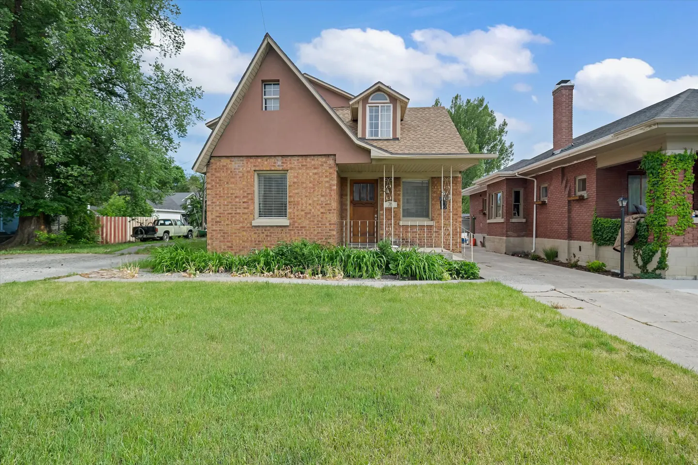 A two-story brick and brown-sided house with a front lawn, dormer window, and a concrete driveway on a sunny day.