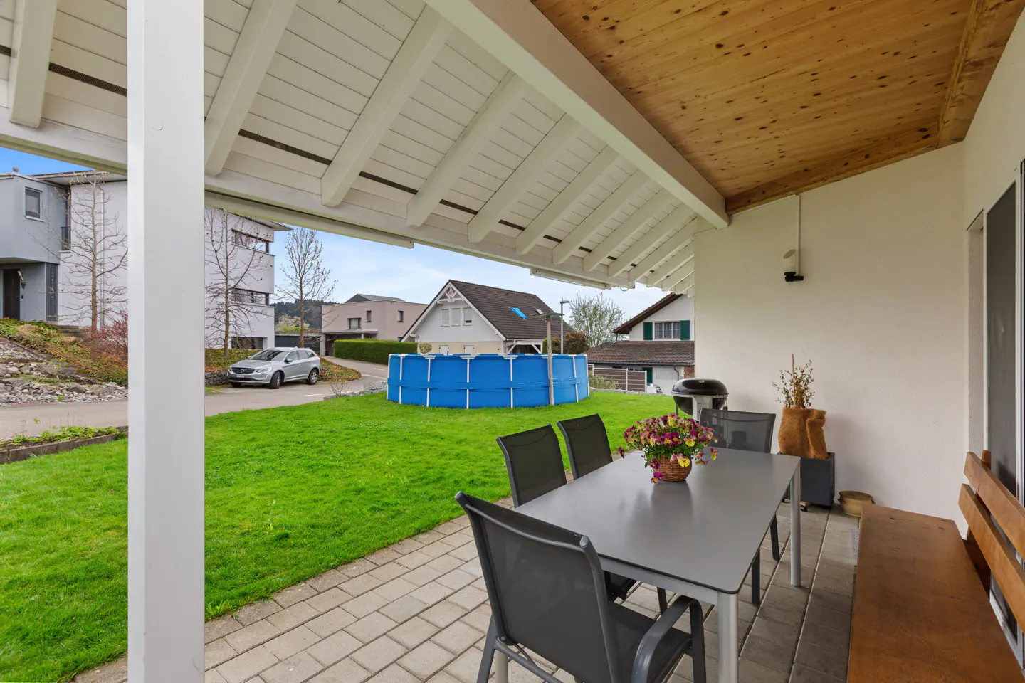 Covered patio with a gray table, black chairs, and a wooden bench. A blue pool is in the green yard, with houses in the background.