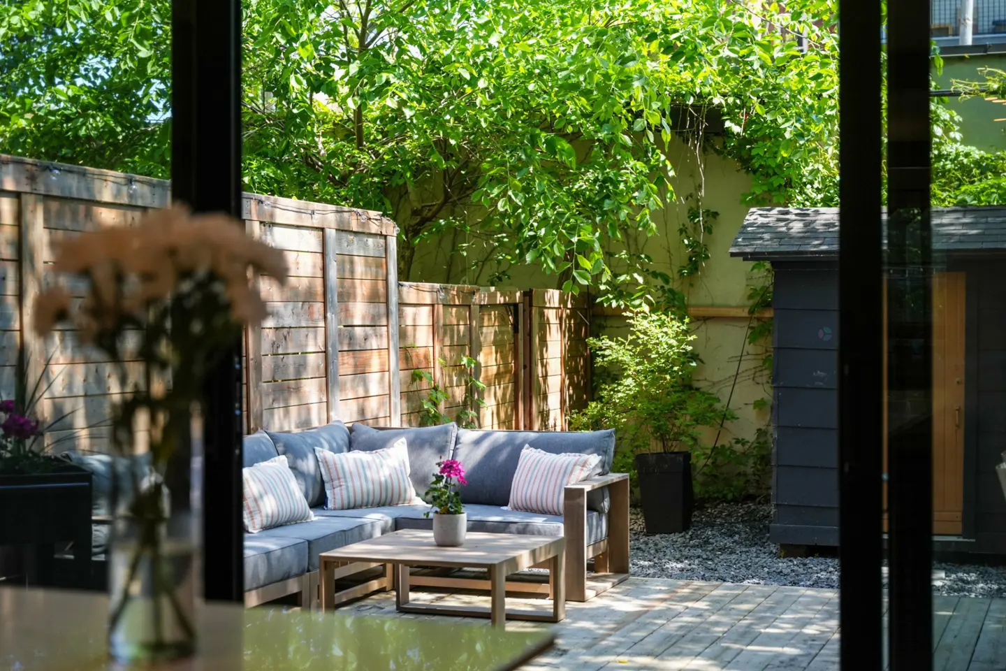 Outdoor patio with gray sectional sofa, wooden coffee table, and potted plant. Wooden fence and green trees in the background.