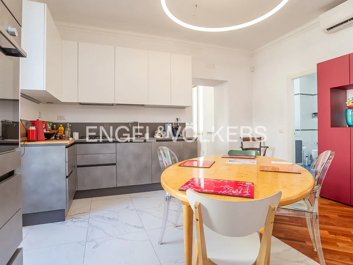 A modern kitchen with white cabinets, gray counters, and a round wooden table with chairs. A red cabinet is visible in the background.