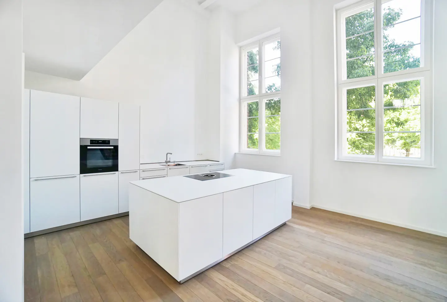 Bright, modern kitchen with white cabinets, island, and hardwood floors. Two large windows offer a view of green trees.