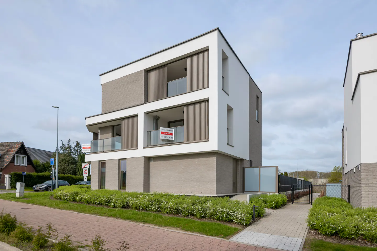 Modern three-story apartment building with white and gray brick exterior, glass balconies, and green landscaping. "For Sale" sign visible.