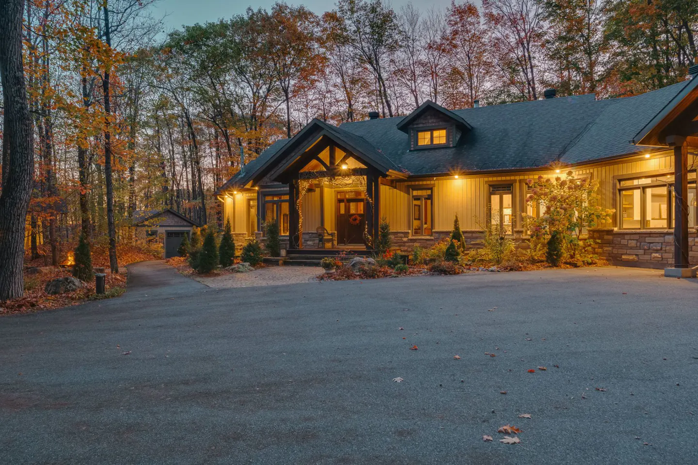 A tan house with a dark roof is surrounded by fall foliage and lit by warm lights.