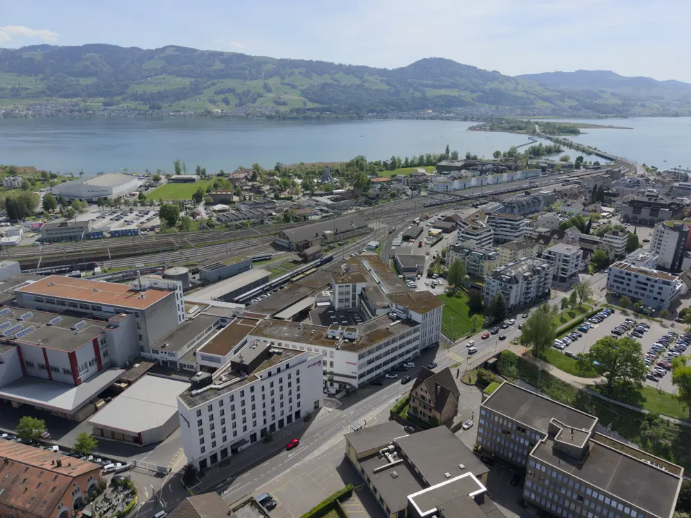Aerial view of Rapperswil, Switzerland, showing buildings, train tracks, and Lake Zurich with mountains in the background.