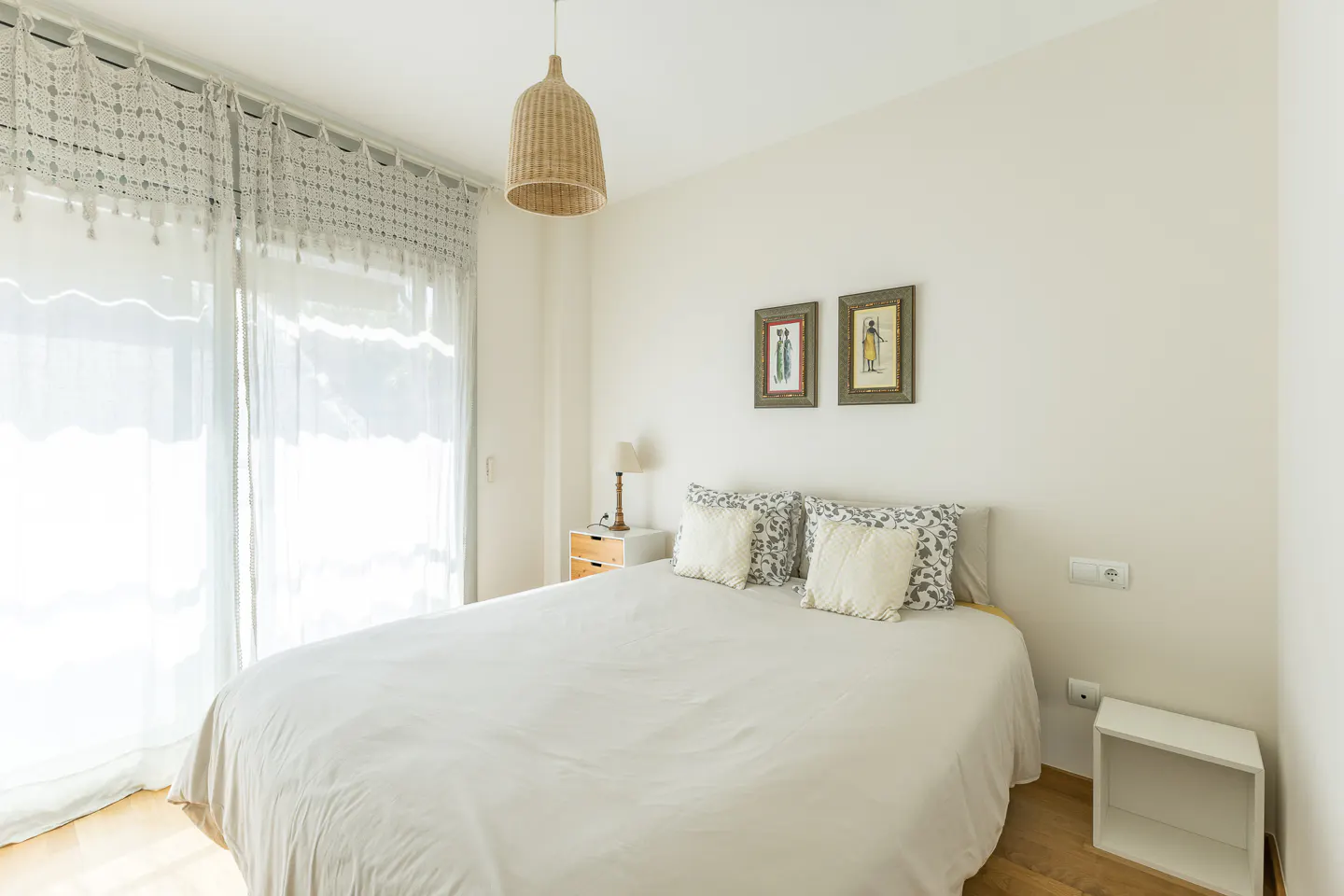 Bright bedroom with a white bed, patterned pillows, and a woven pendant light. Two framed pictures hang on the wall above the bed. Curtains cover the window.