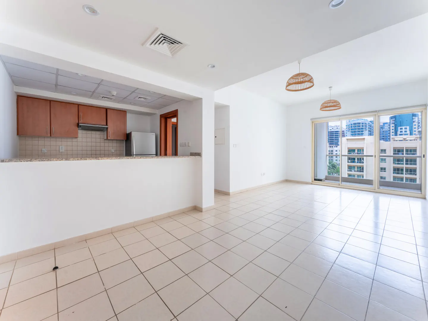 Bright, empty apartment with white walls, tile floors, and an open kitchen. A balcony offers a city view. Two wicker pendant lights hang from the ceiling.