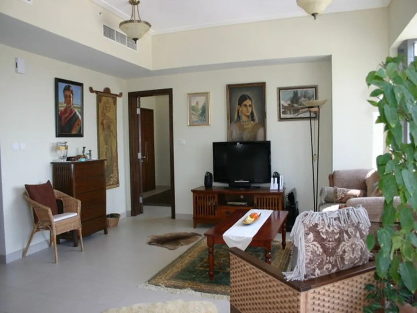 Living room with beige walls, tile floors, and a mix of traditional furniture. Paintings adorn the walls above a TV and wooden dresser.