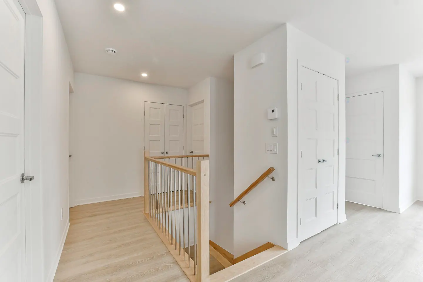 Bright hallway with white walls, doors, and light wood floors. A wooden railing overlooks the stairs.