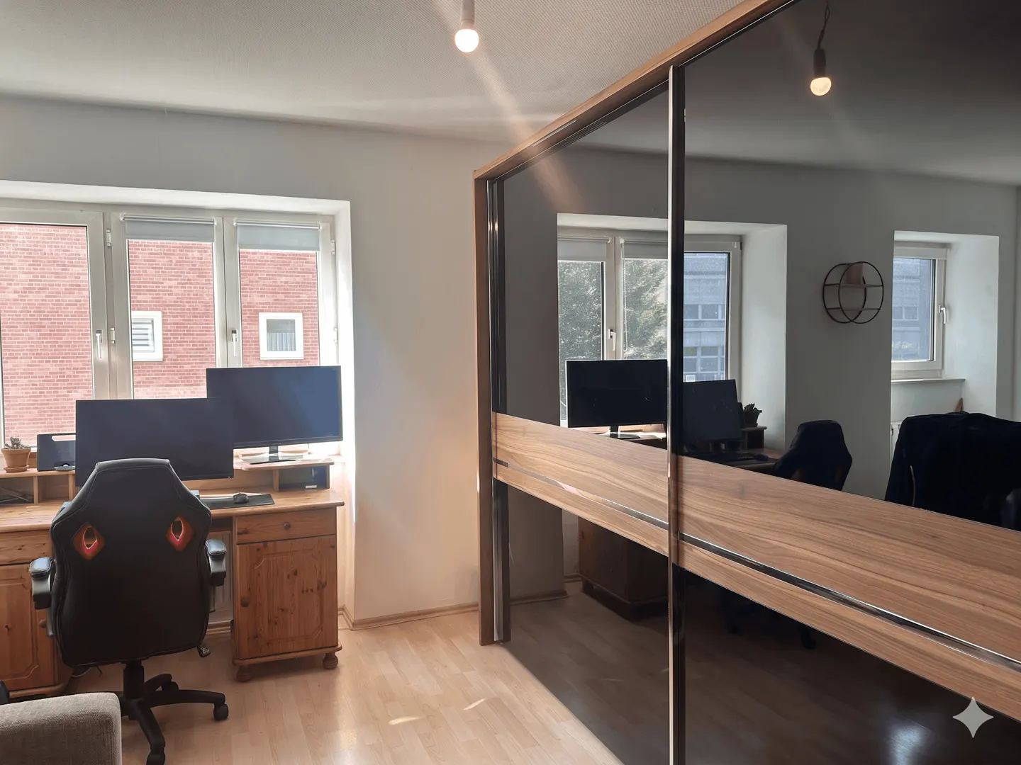 Home office with a wooden desk, dual monitors, and a gaming chair, featuring a large mirrored sliding door closet.