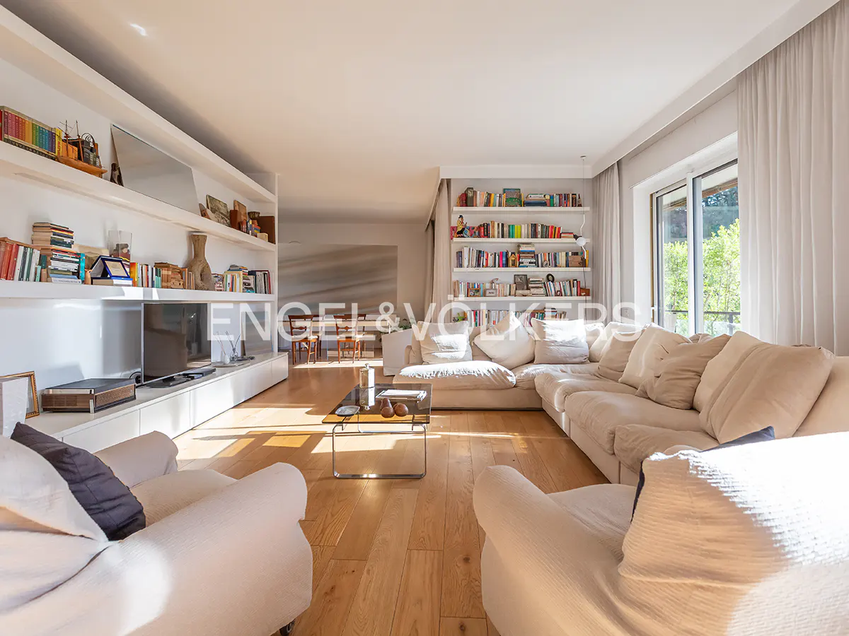 Bright living room with hardwood floors, white sofas, bookshelves, and a glass coffee table. Natural light streams in from a window.