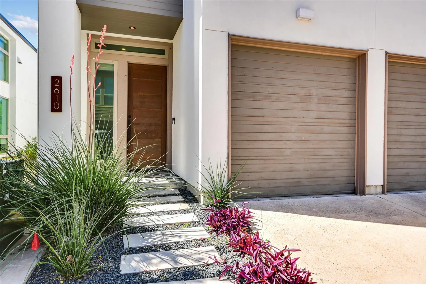 Modern home exterior with a brown door, two-car garage, stone walkway, and green and purple landscaping.
