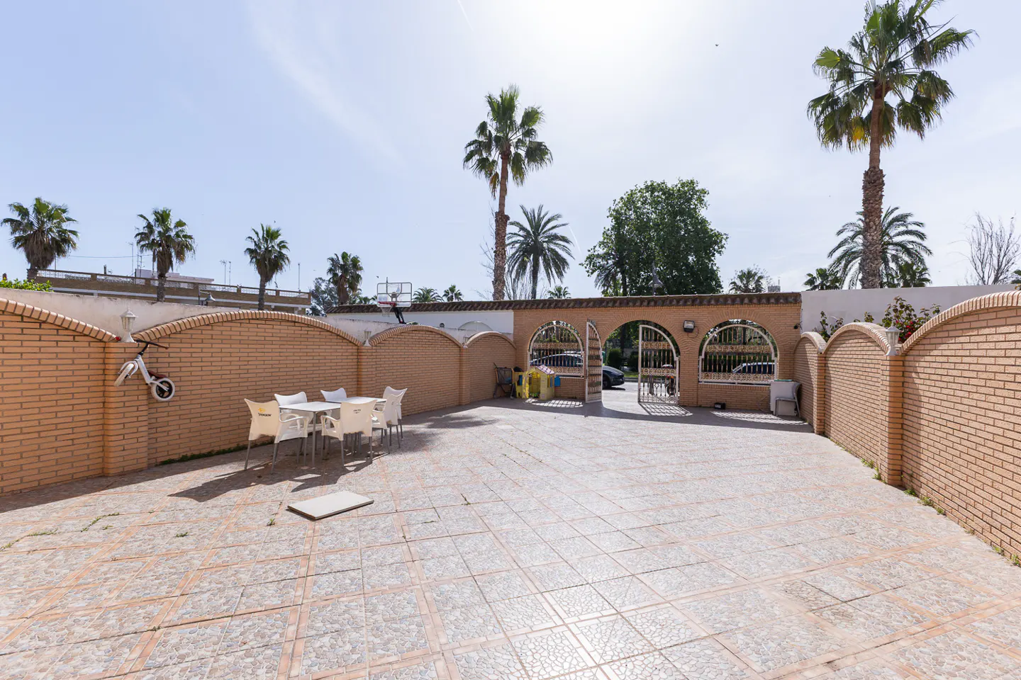 Outdoor patio with tiled floor, brick walls, and palm trees. A white table and chairs sit near a wall with a bicycle hanging on it.