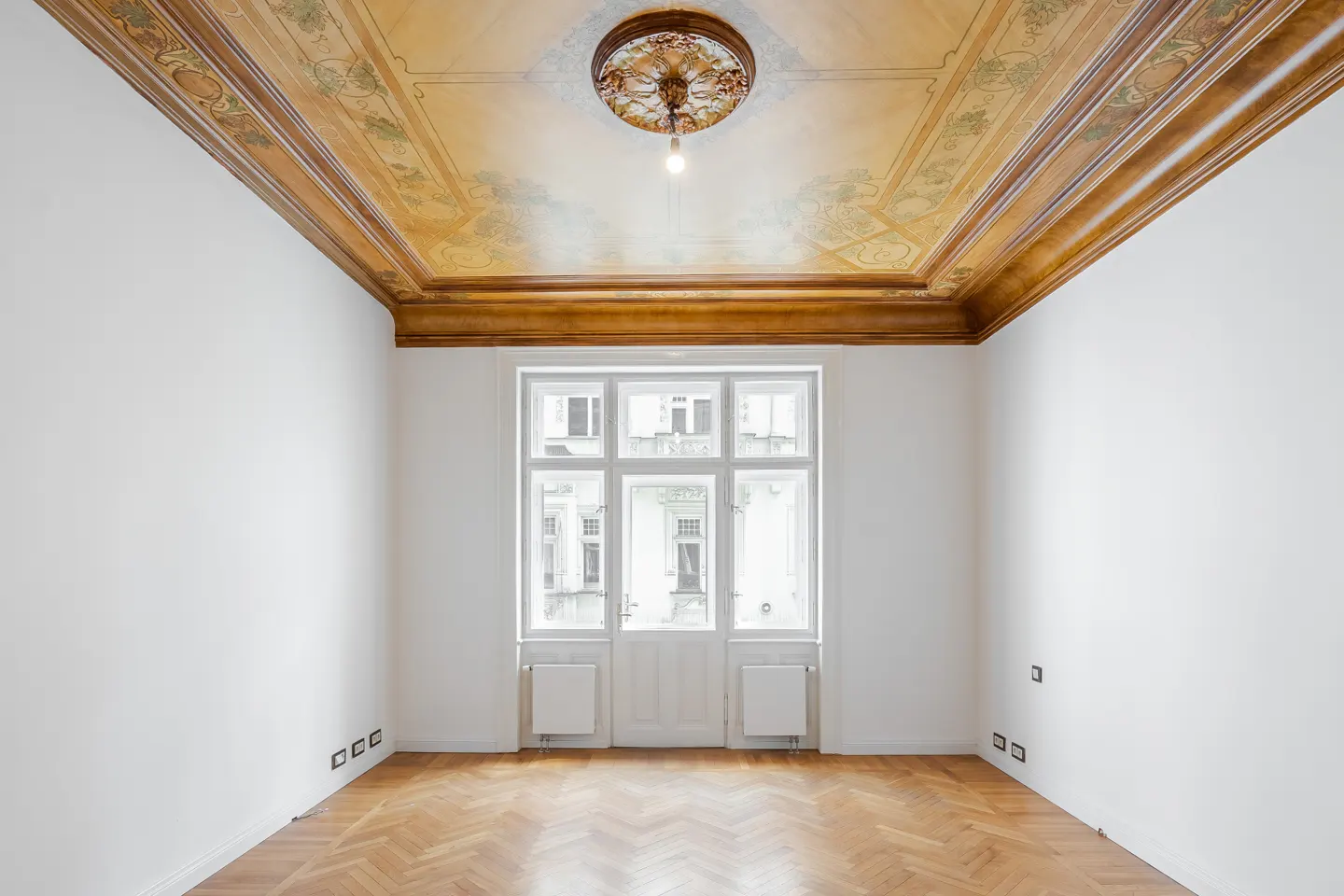 Bright, empty room with white walls, herringbone wood floor, and ornate ceiling. A large window/door lets in natural light.