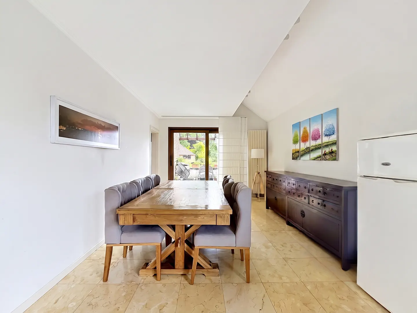 A dining room with a long wooden table, gray chairs, a dark wood buffet, and a white refrigerator.