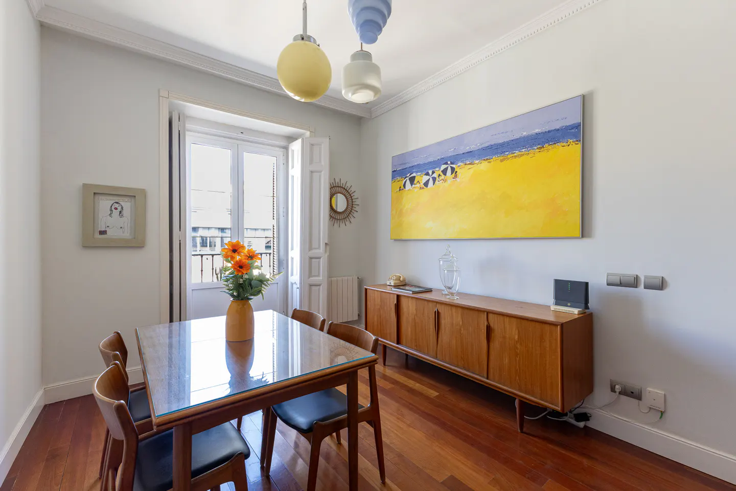Dining room with glass table, wooden chairs, mid-century sideboard, and a large beach painting on a white wall.
