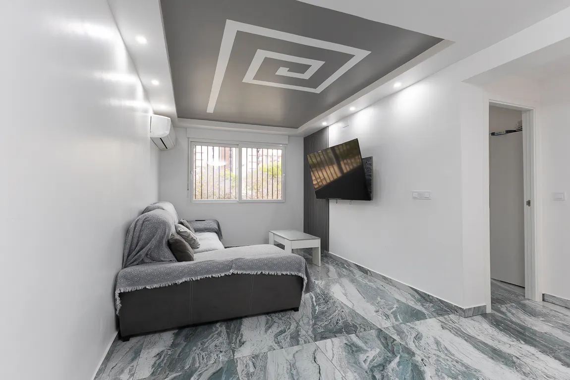 Living room with gray sofa, white walls, and marble floor. A TV hangs on the wall, and a window provides natural light. The ceiling has a gray and white geometric design.