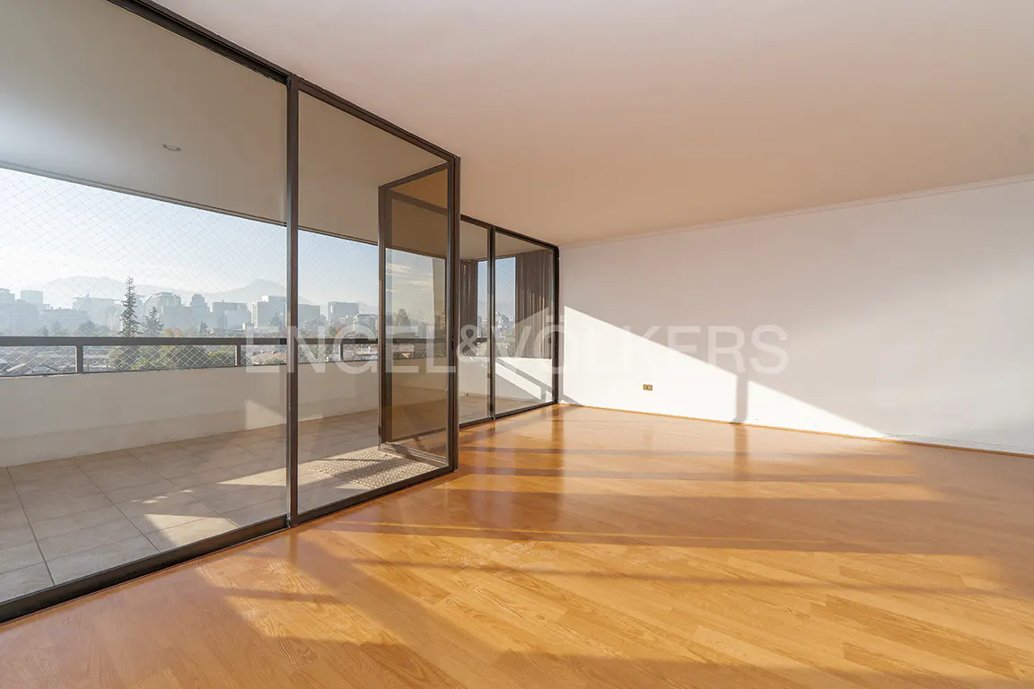 Bright, empty room with light wood floors and white walls. Large glass doors open to a balcony with a city view.