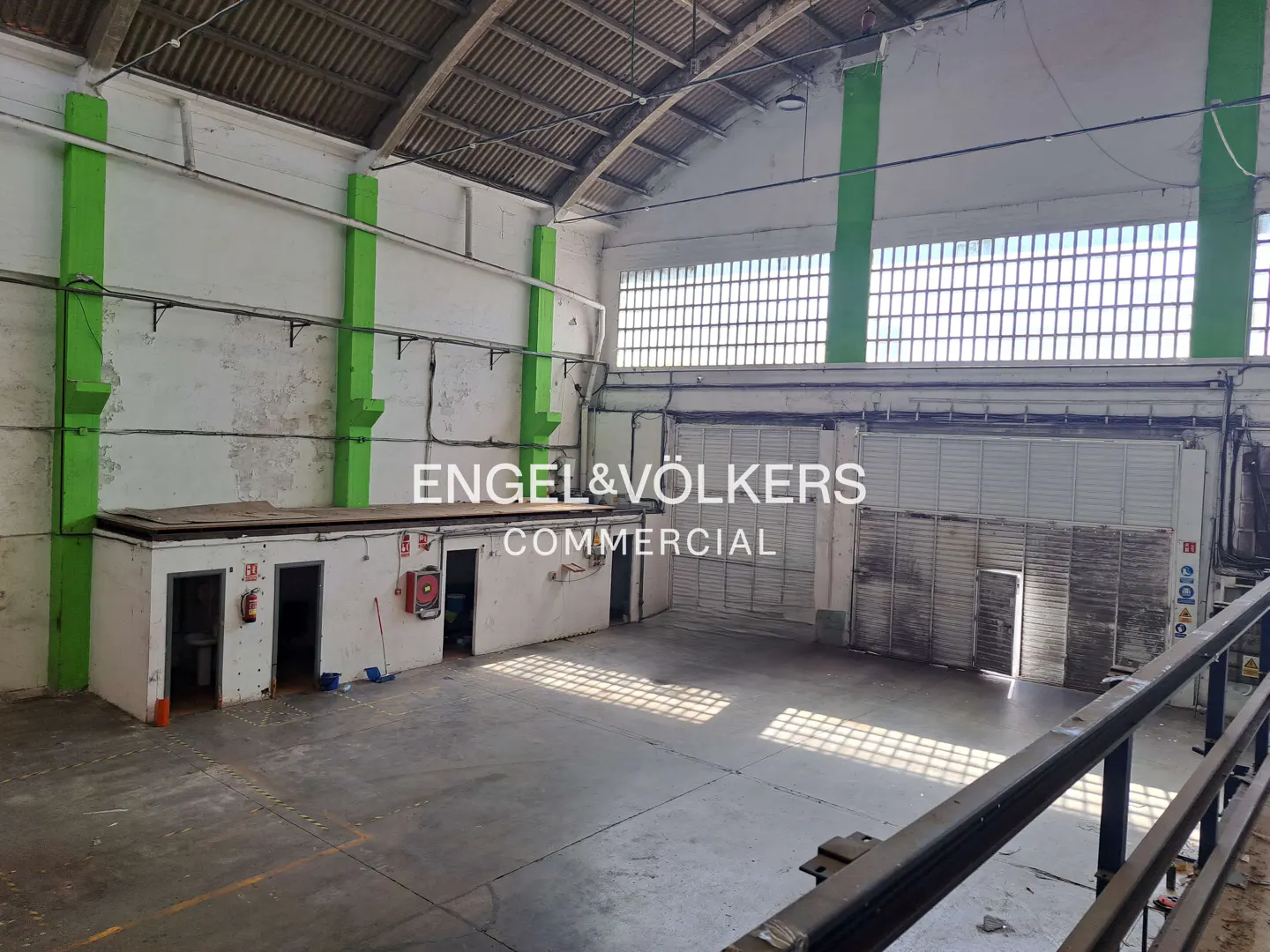 A large, empty warehouse with white walls, green pillars, and a high, arched ceiling. Windows let in sunlight, creating shadows on the gray floor. "Engel & Völkers Commercial" is superimposed.