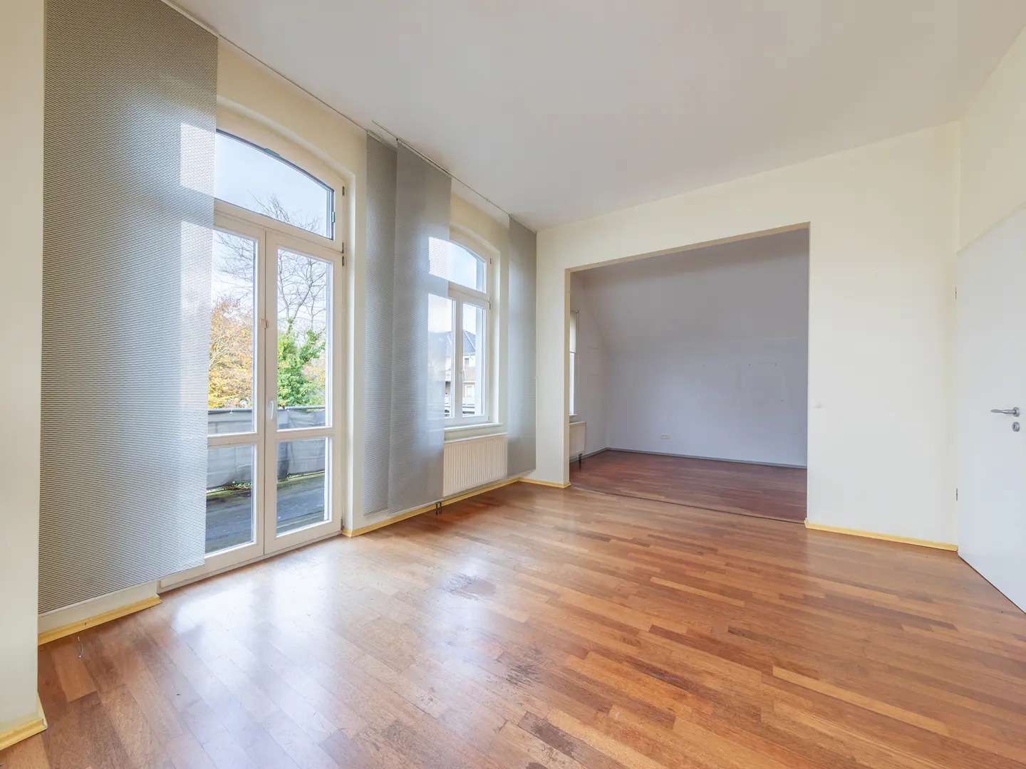 Empty room with hardwood floors, white walls, and large windows with gray curtains. An open doorway leads to another room.