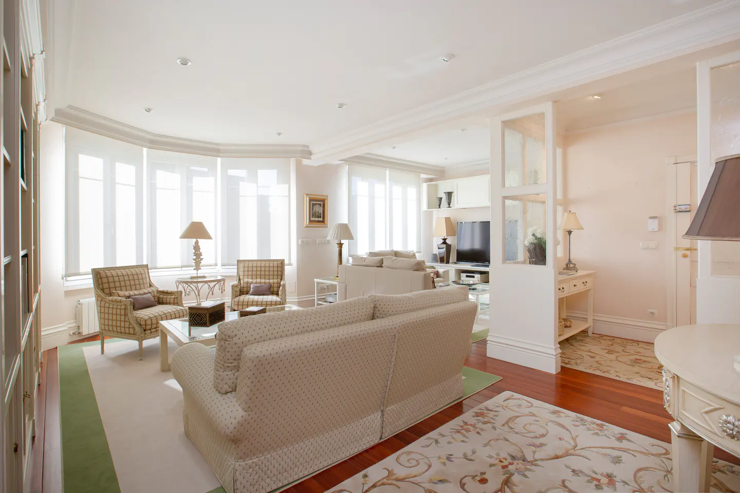 Bright living room with beige sofa, plaid armchairs, and large windows. Hardwood floors and floral rug add warmth.