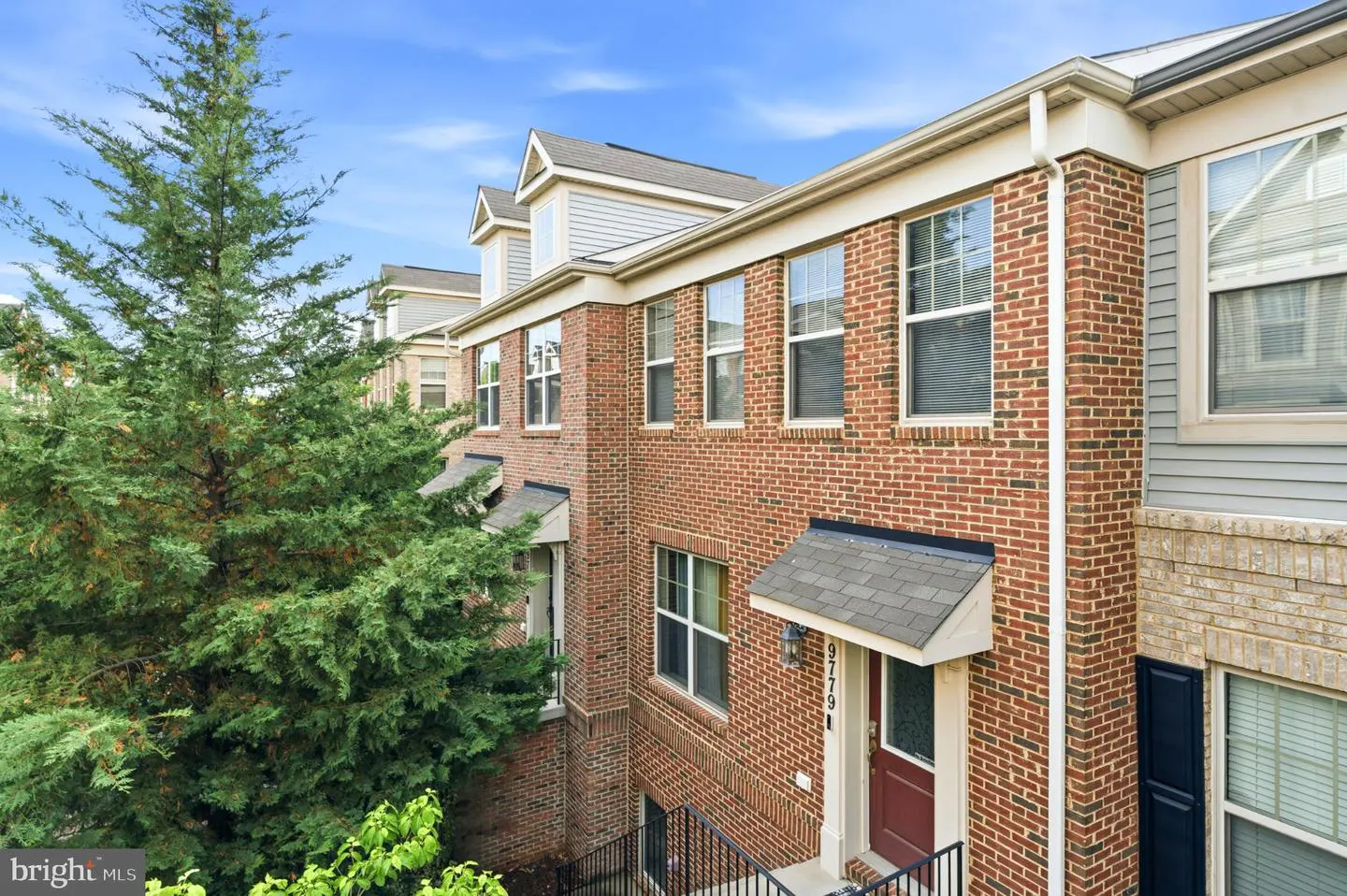 Exterior view of a multi-story red brick townhouse with a dark front door, white trim, and a large evergreen tree nearby.