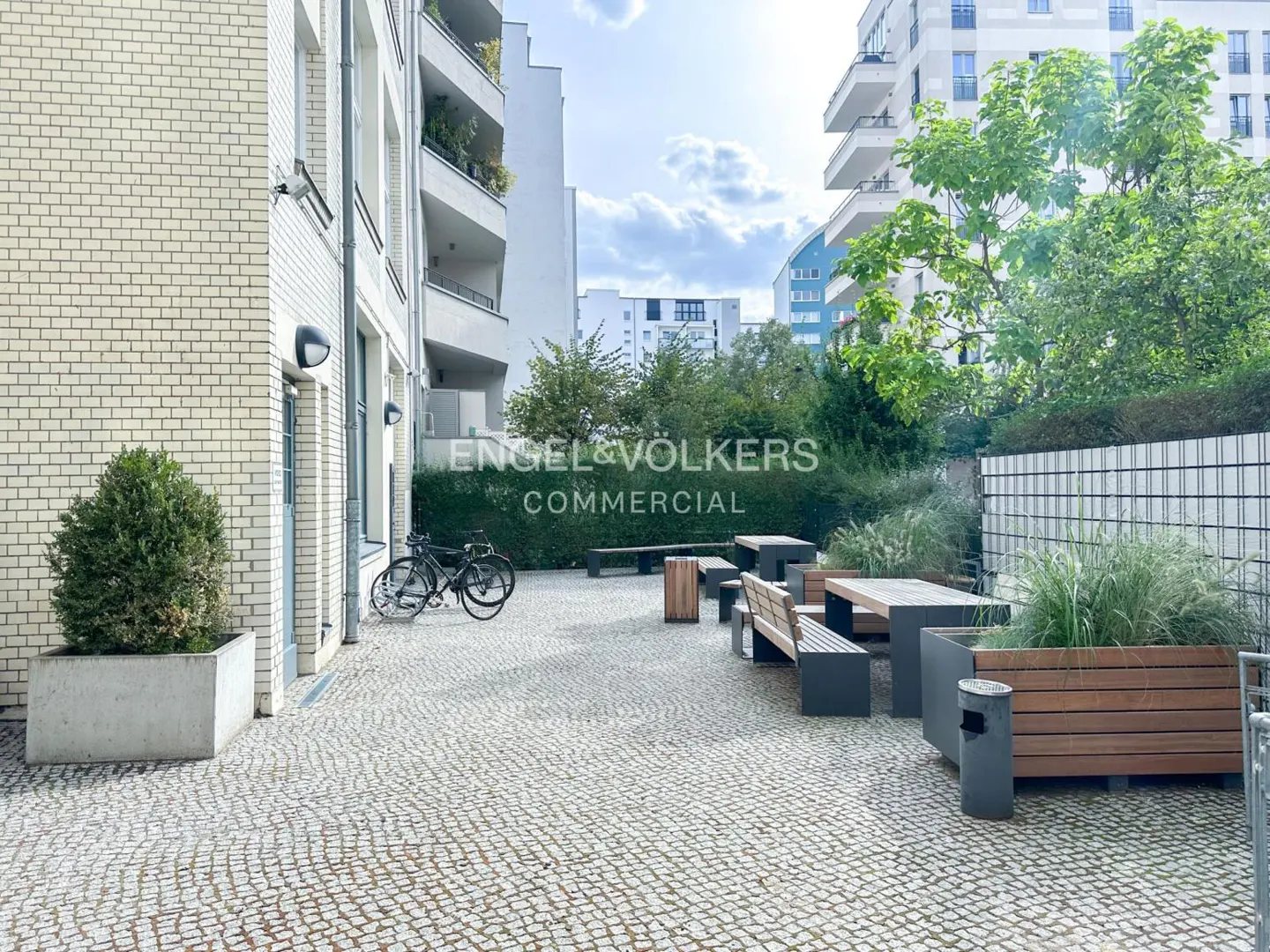 Outdoor courtyard with stone pavers, benches, tables, and greenery. Two bicycles are parked near a white brick building.