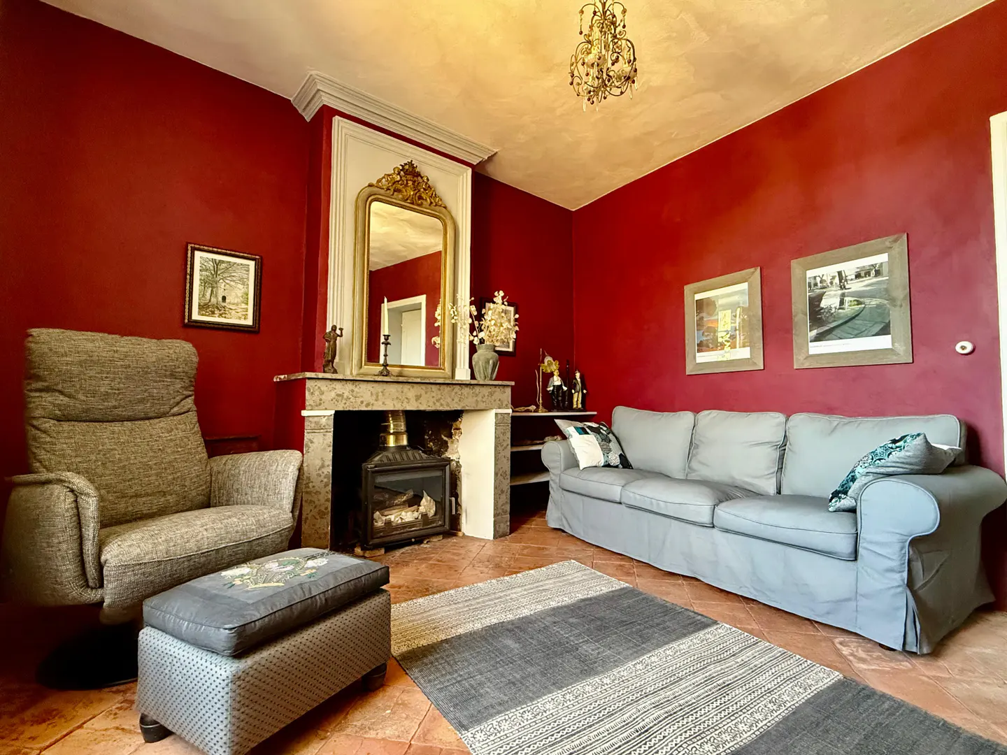 Living room with red walls, gray sofa and chair, fireplace with gold mirror, and gray rug on terracotta tile floor.