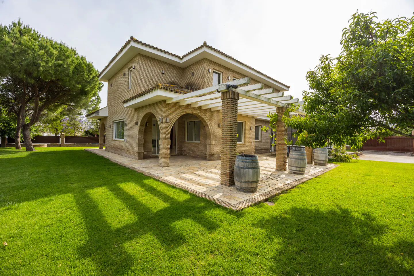 Exterior view of a two-story brick house with a pergola, patio, and green lawn. Three wooden barrels decorate the patio.