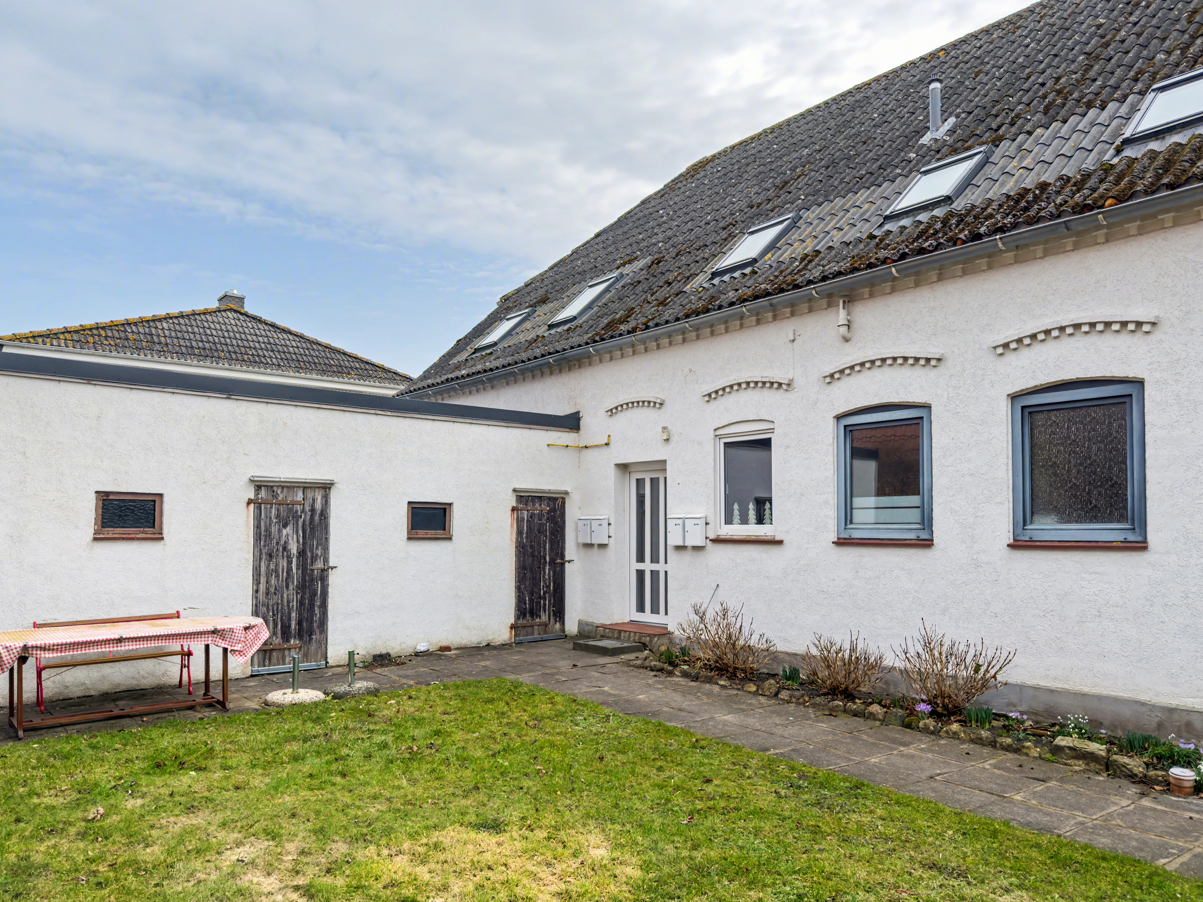 Exterior view of a white building with a gray tiled roof and skylights, a green lawn, and a table with a red and white checkered tablecloth. [AI-generated text]
