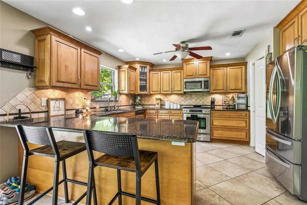 A kitchen with wood cabinets, granite countertops, tile backsplash, and stainless steel appliances. Two bar stools sit at the island.