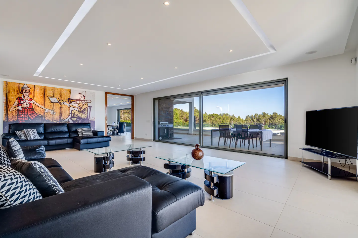 Bright living room with black leather sectional sofa, glass coffee tables, and a large painting. Sliding glass doors lead to an outdoor dining area.
