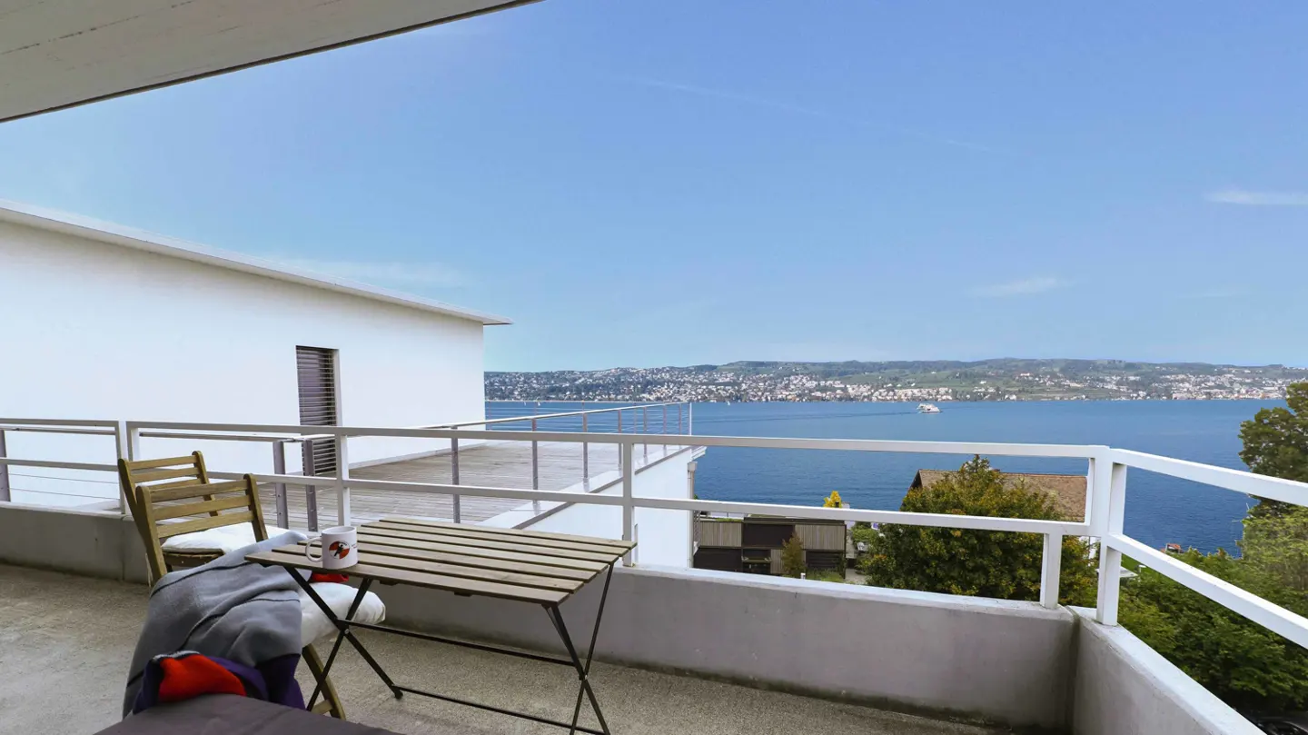 Balcony view of a lake. White building with a table, chair, blanket, and mug on the balcony. Blue sky and water.