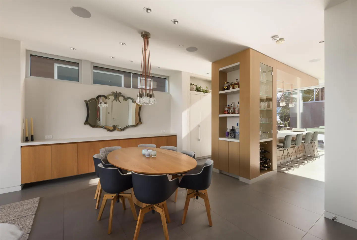 Bright dining room with an oval wood table, gray chairs, and a modern chandelier. Built-in cabinets and a bar area are in the background.