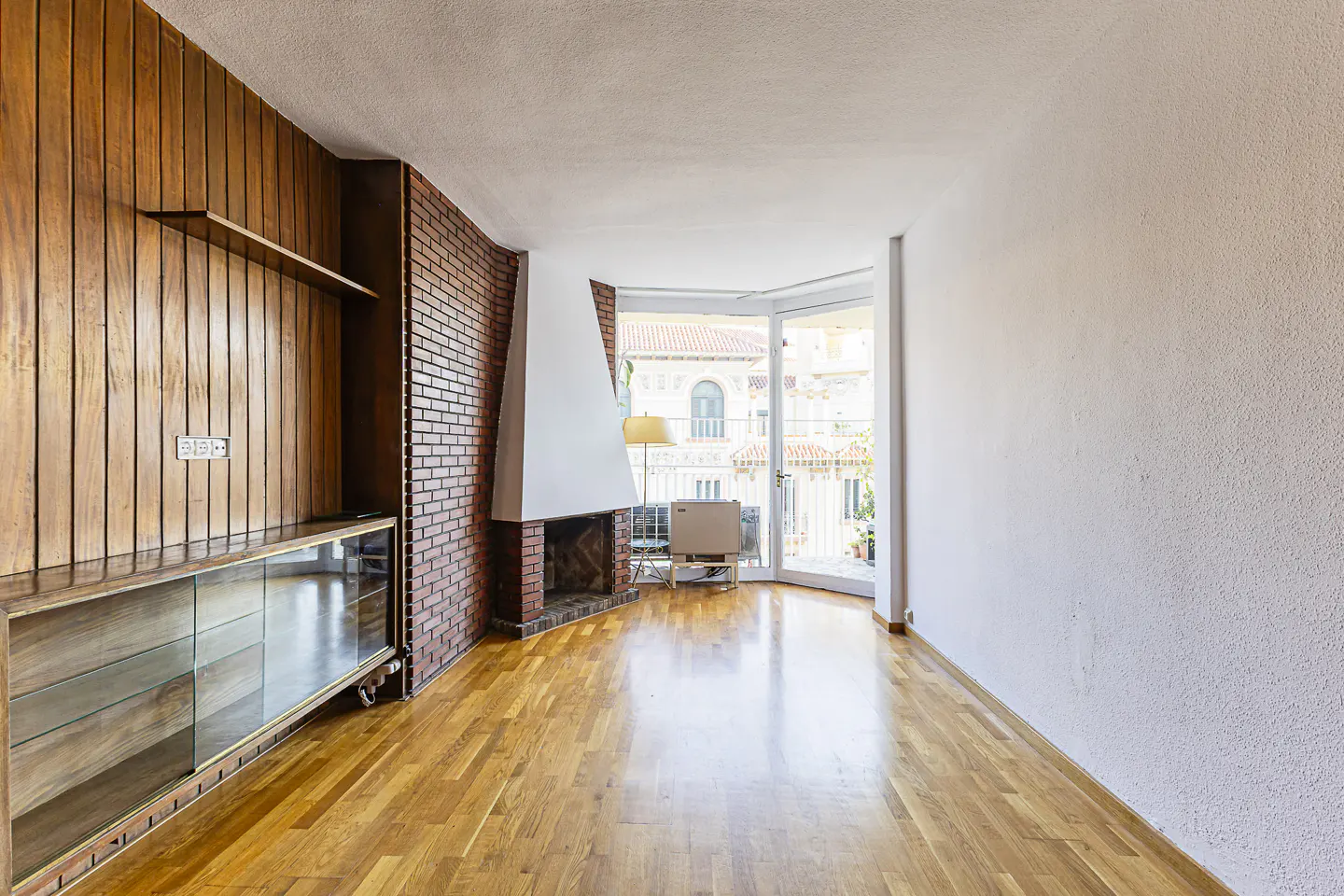 Bright living room with wood floors, brick fireplace, and wood paneled wall with glass cabinet. Balcony with view of buildings.