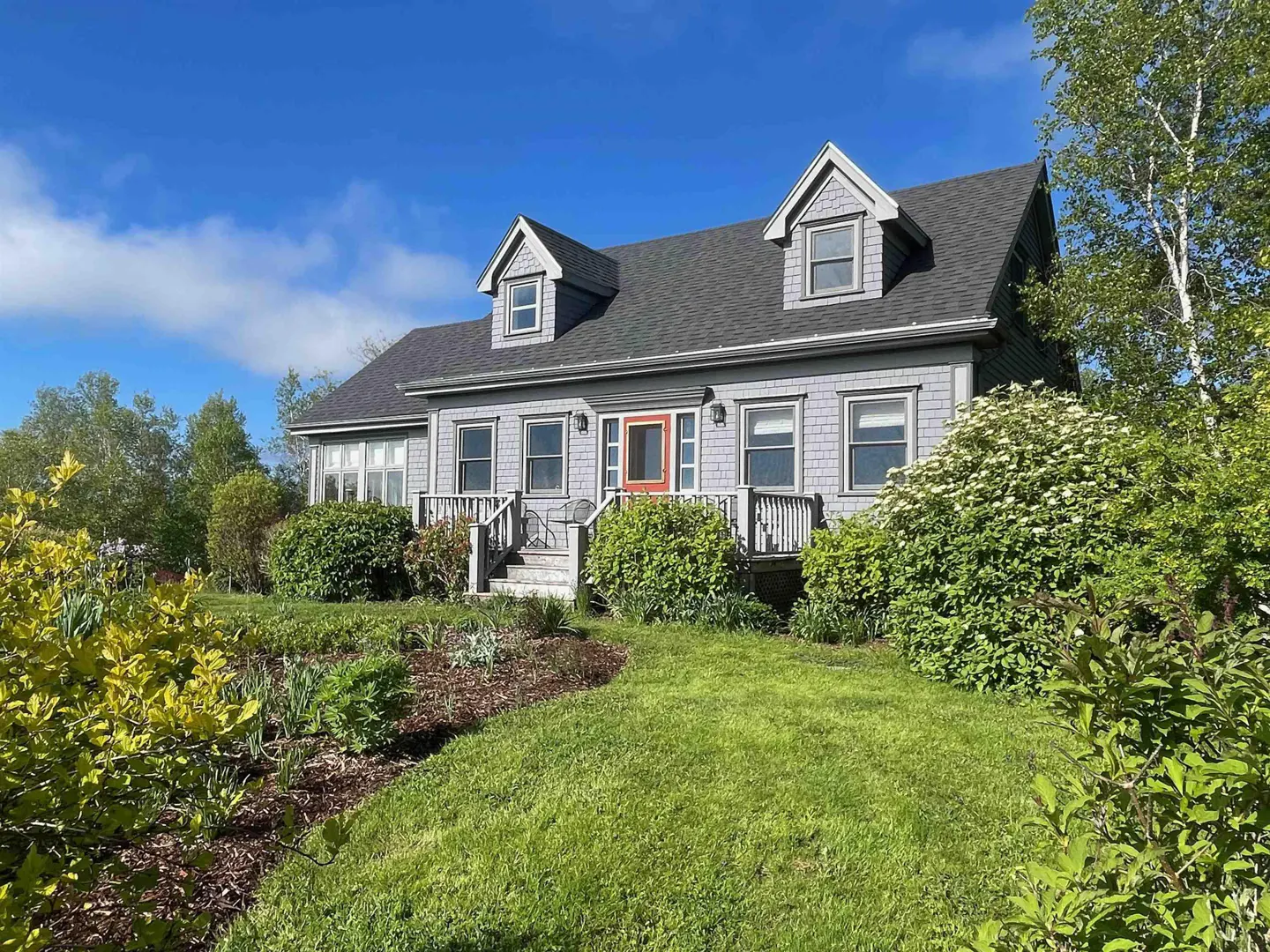 A grey shingled house with a bright orange front door, dormer windows, and a front porch, set in a lush green yard.