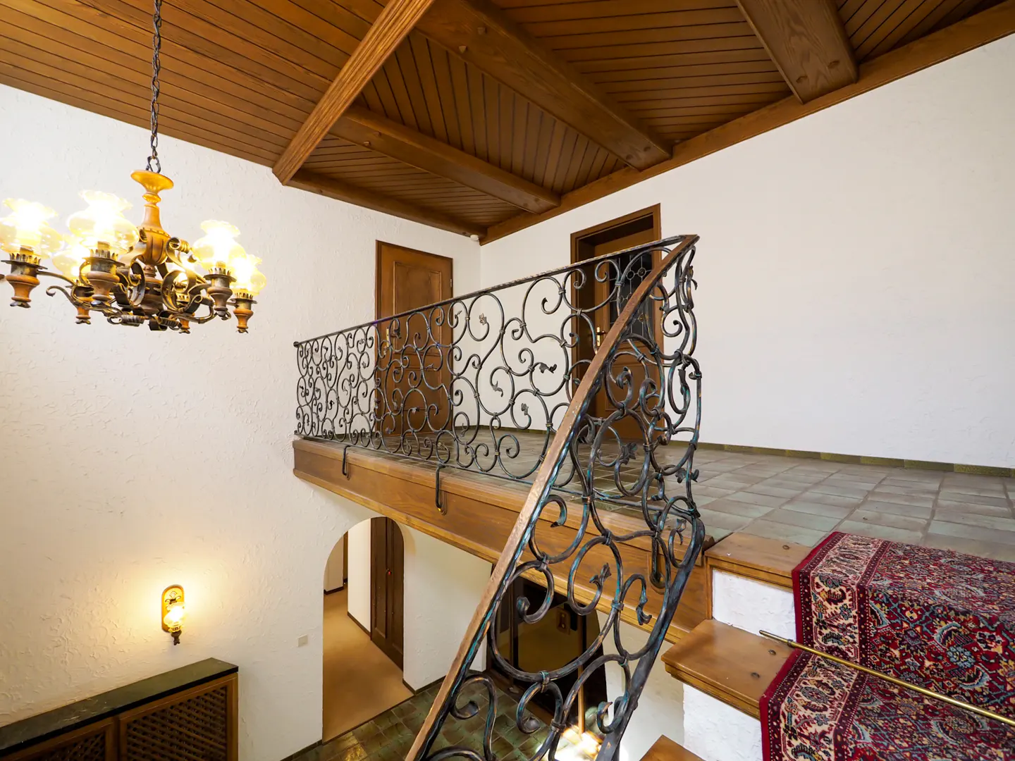 Interior view of a home's foyer with a wooden ceiling, chandelier, and staircase with a decorative wrought iron railing.