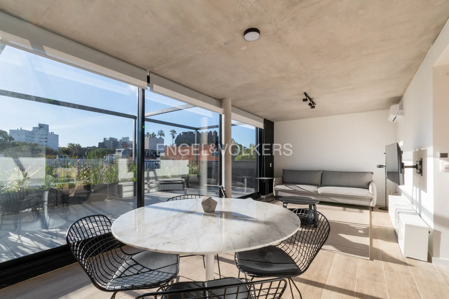 Bright apartment interior with a round marble table, black wire chairs, and a gray sofa. Large windows offer a city view.