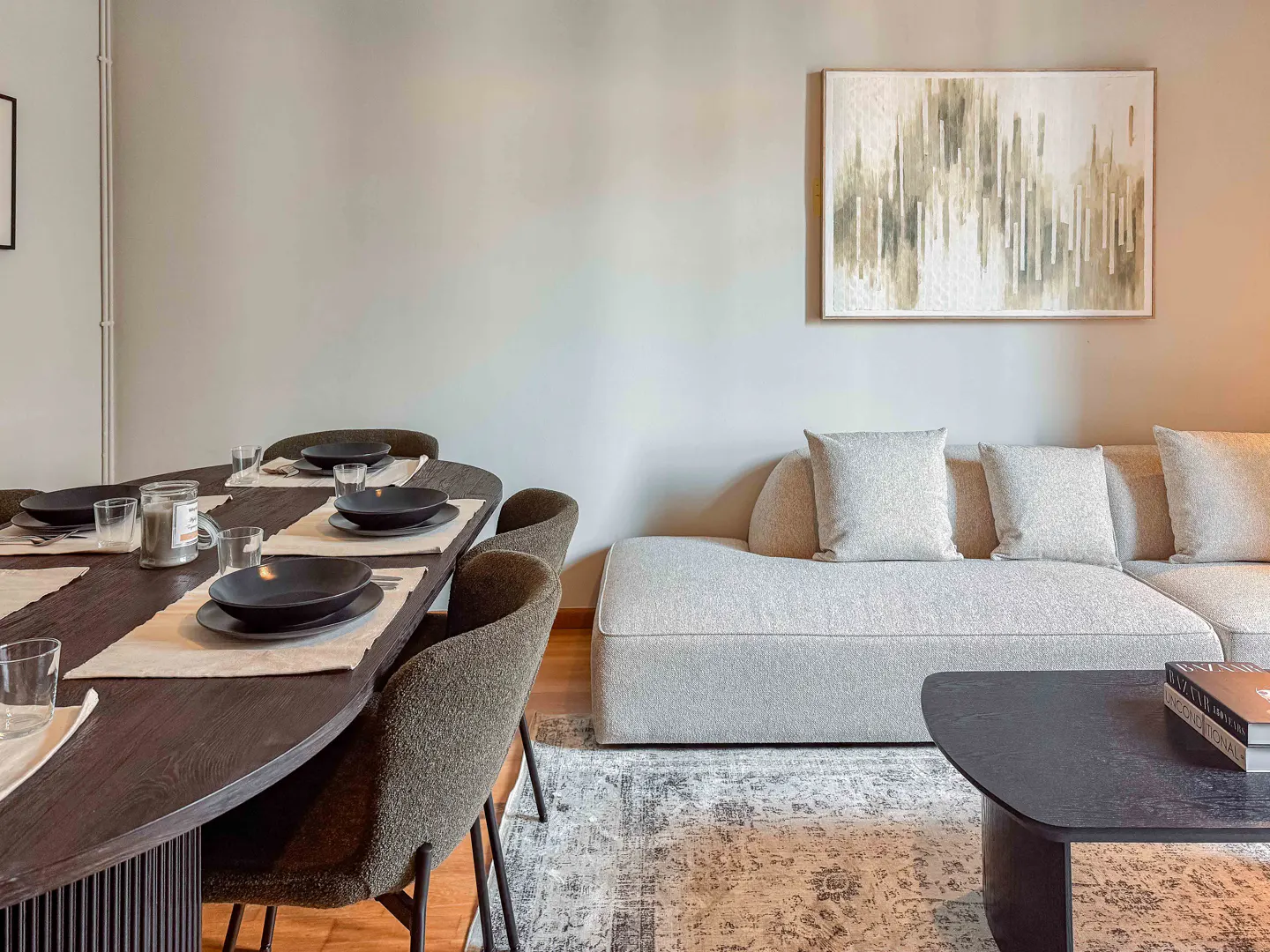 Living room with a dark wood dining table set for a meal, a light gray sofa, and a patterned rug. An abstract painting hangs on the wall.