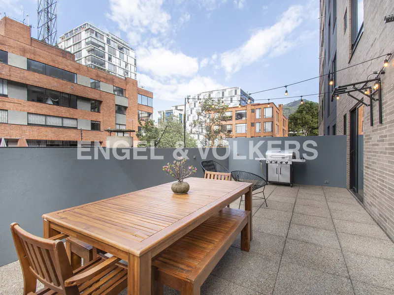 Outdoor patio with a wooden table, bench, chairs, and grill. Buildings are visible in the background.