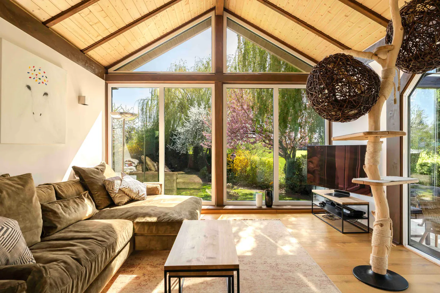 Bright living room with wood ceiling, large windows overlooking a garden, brown sofa, and a cat tree.