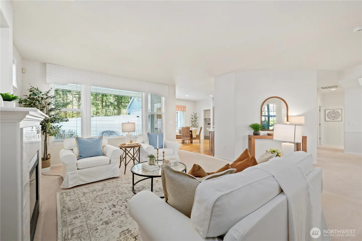 Bright living room with white sofas, blue pillows, and a round glass coffee table on a patterned rug. Large windows offer natural light.