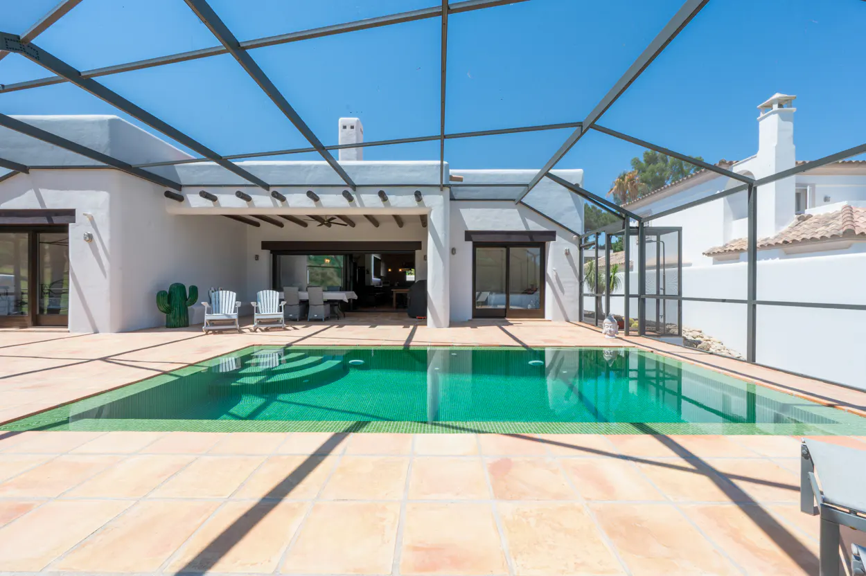 A screened-in patio with a green swimming pool, tan tile deck, and white house exterior under a clear blue sky.