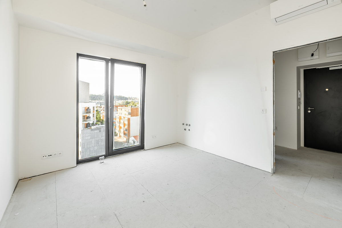 Empty, unfurnished room with white walls, grey flooring, a glass balcony door, and a black door leading to a hallway. [AI-generated text]