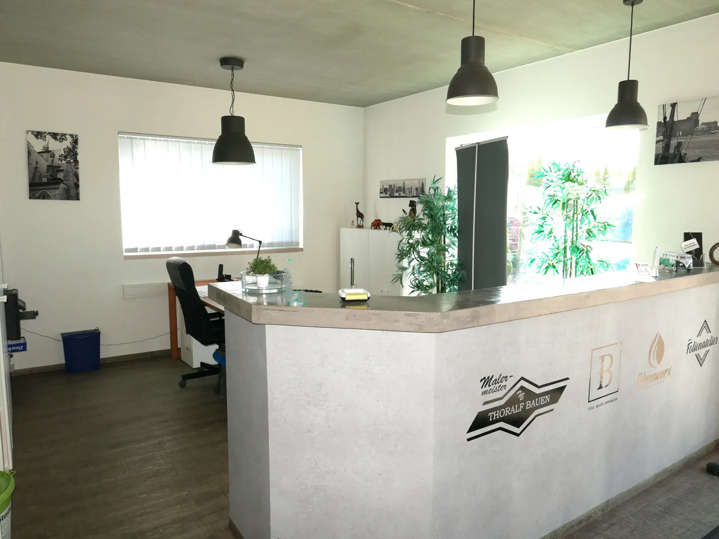 Modern office reception area with a concrete-style desk, black pendant lights, indoor plants, and a desk chair.